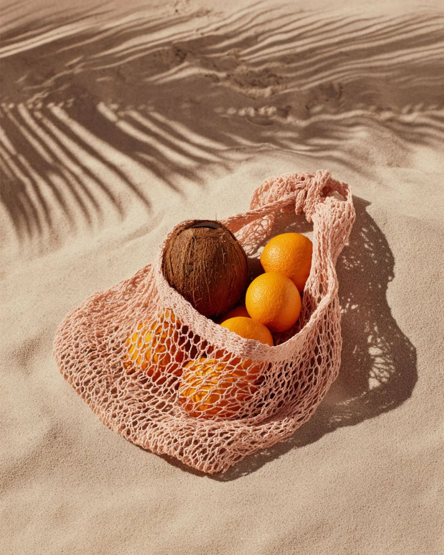 Reusable mesh bag with coconut and oranges on sandy beach with palm leaf shadows