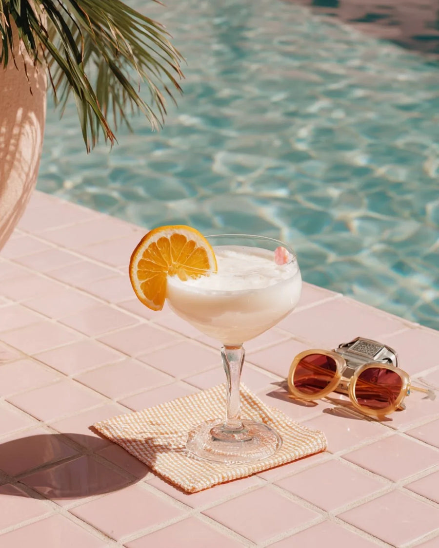 Refreshing poolside drink with orange slice, sunglasses, and palm leaves on pink tiles