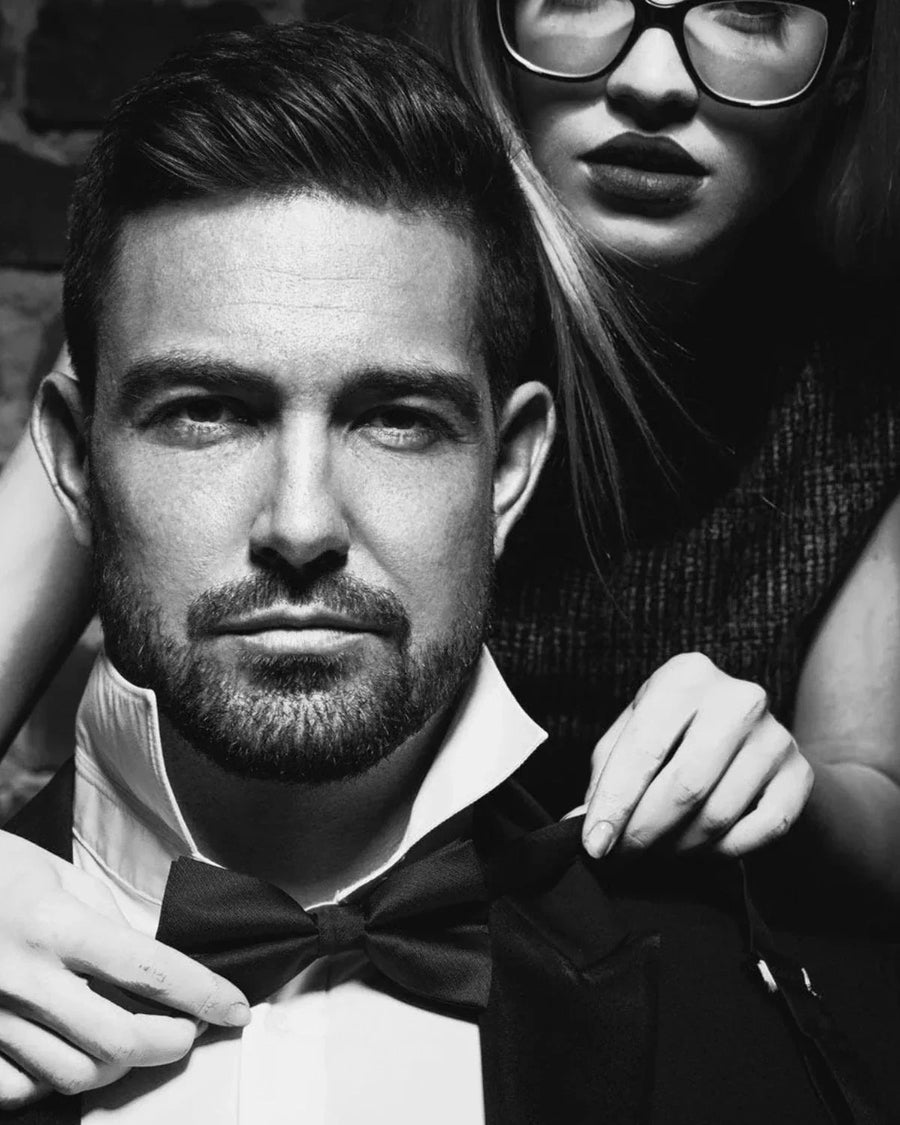 Black and white portrait of a man in formal wear adjusting a bow tie with a woman behind him.