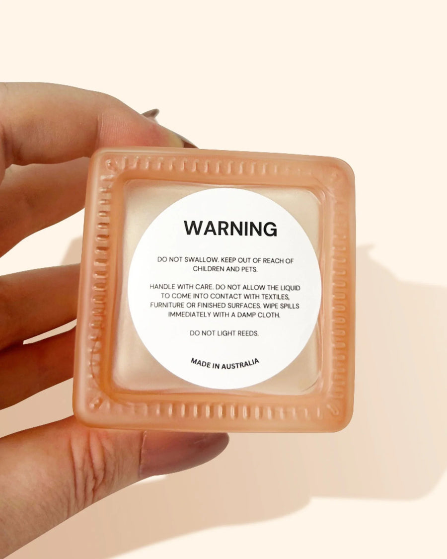 Hand holding square fragrance oil bottle with safety warning label, peach cap shown.