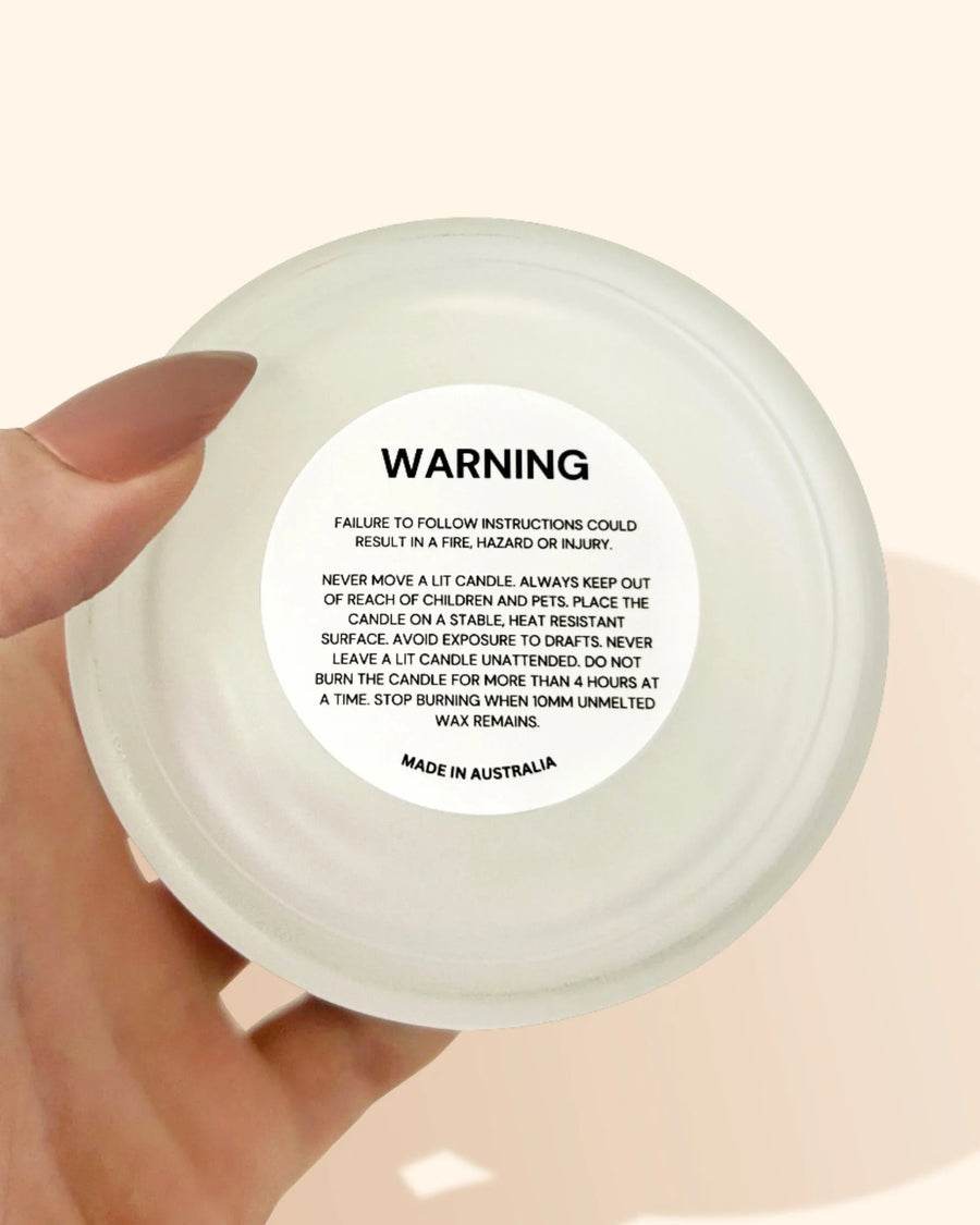 Hand holding a candle jar with warning label and safety instructions on the lid.