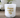 White glass candle with gold 'happy mother's day' text on wooden table, flowers in background