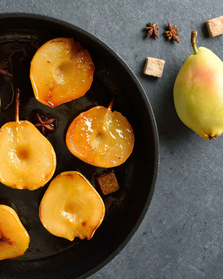Sliced caramelized pears in a pan with star anise, whole pear and sugar cubes on gray surface