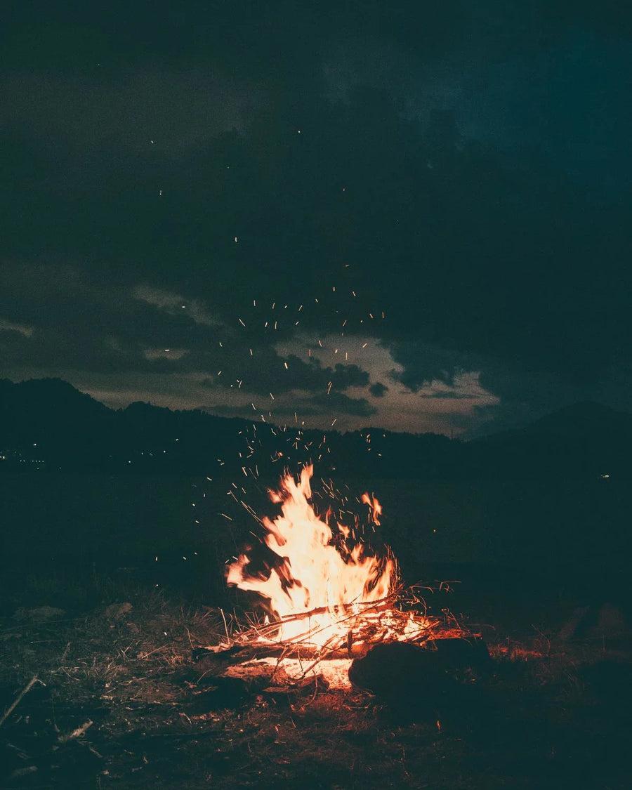 Outdoor campfire burning with sparks under a dark, cloudy night sky.