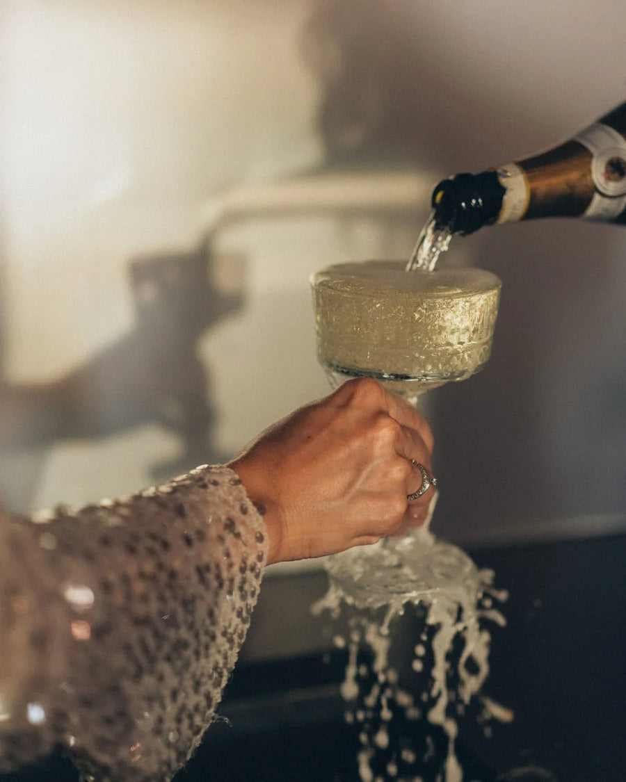 Woman in sequined sleeve holding a glass as sparkling wine overflows while being poured