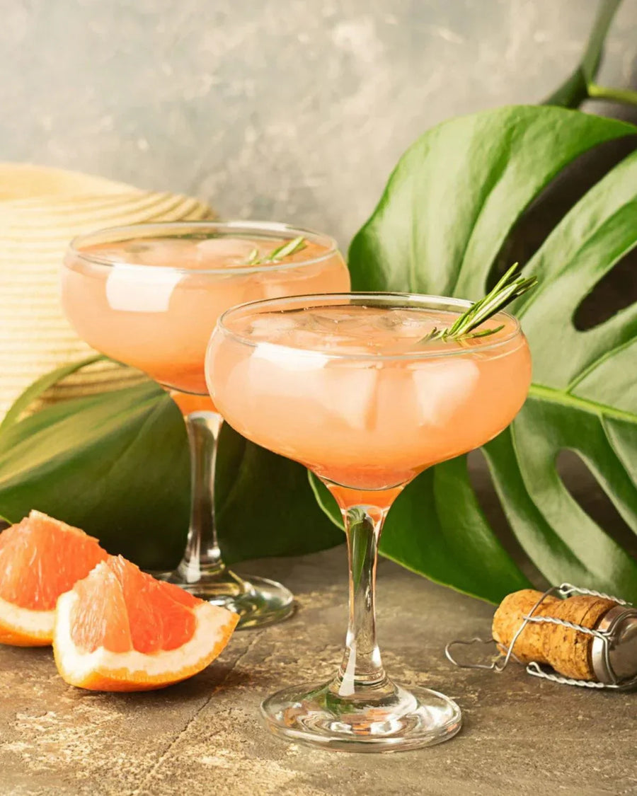 Two grapefruit cocktails with ice and rosemary garnish in coupe glasses, next to grapefruit wedges, on a stone surface with green leaves and a cork nearby.