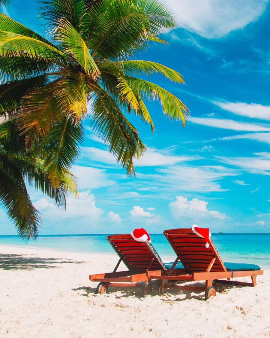 Two beach chairs with Santa hats under palm tree on tropical sandy beach, blue sky