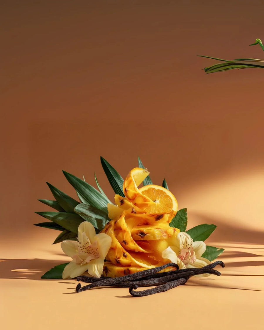 Sliced pineapple, orange, vanilla beans, and flowers arranged on a tan background