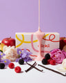 Assorted fruits, vanilla pods, and flowers with pink wax pour and a scented candle box