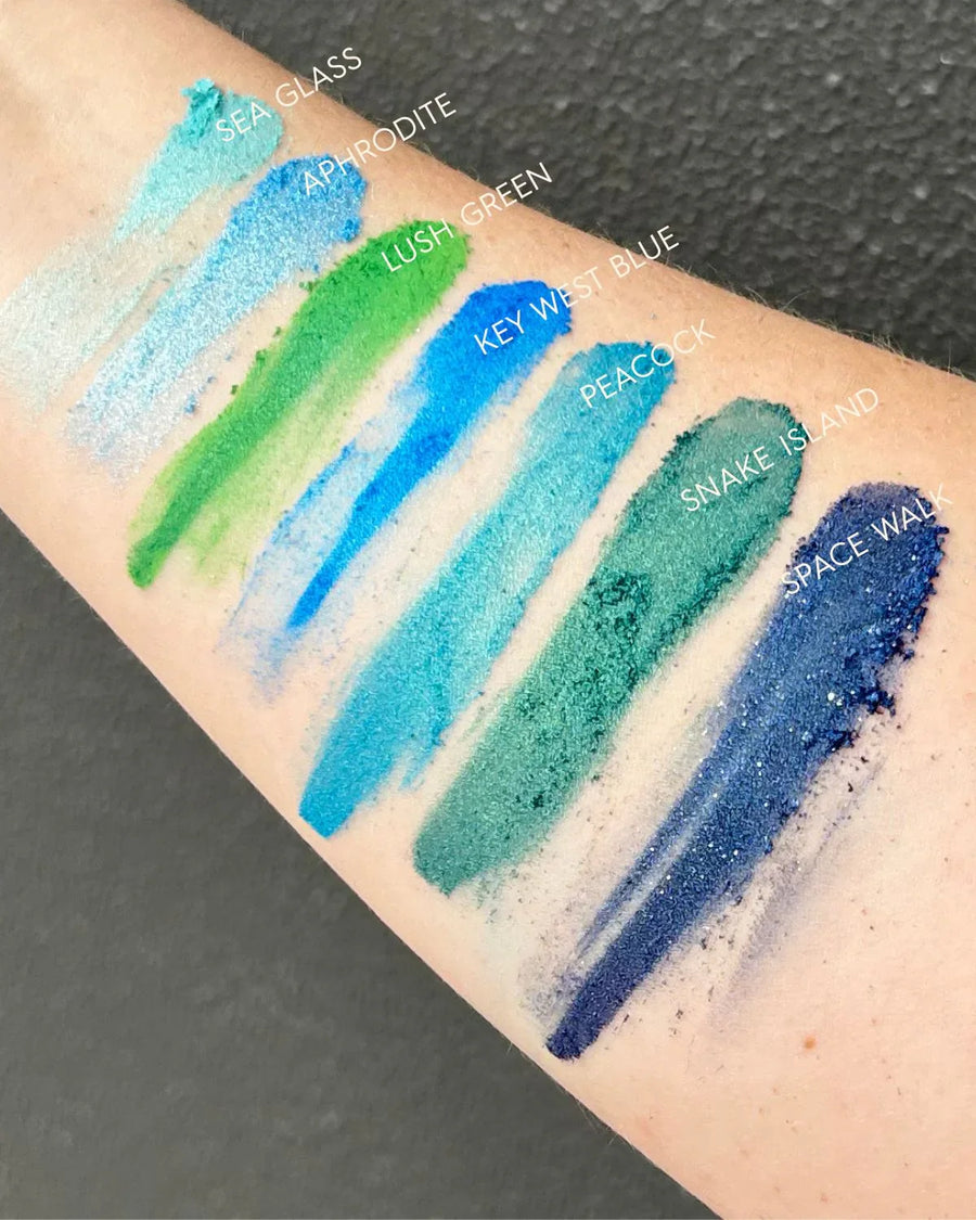 Arm swatched with various blue and green pigment powders labeled Sea Glass, Aphrodite, Lush Green, Key West Blue, Peacock, Snake Island, and Space Walk for soap or candle making.