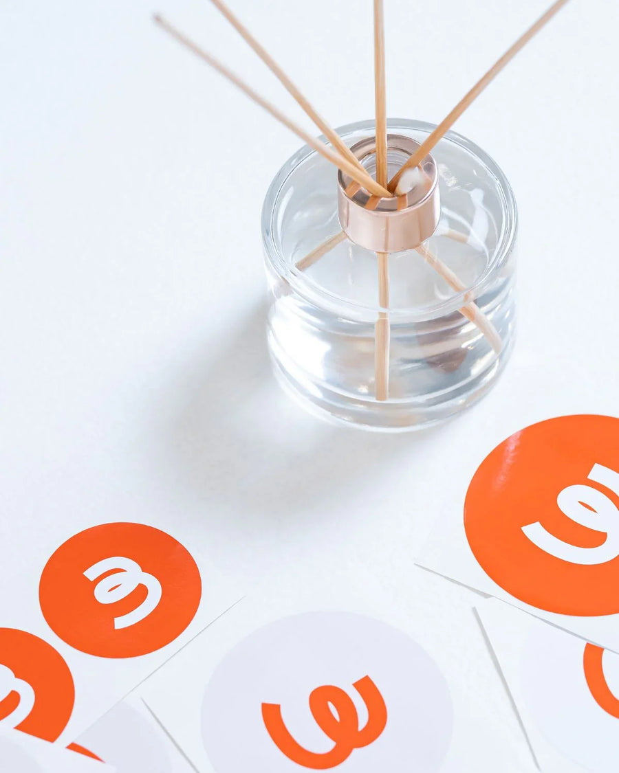 Clear glass reed diffuser with wood sticks and orange logo stickers on white surface